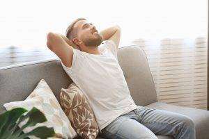 Bearded guy wearing blank white t-shirt & denim pants sitting alone at home on grey textile couch. Young man w/ facial hair in domestic situations. Interior background, copy space, close up, monstera.