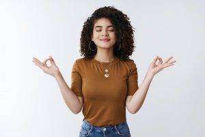 Om girl feels peace patience. Attractive carefree relaxed happy young woman curly shirt hairstyle close eyes smiling delighted meditating hands sideways nirvana lotus pose, breathing practice yoga.