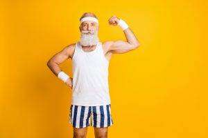 Photo of cheerful positive attractive handsome old man demonstrating, his muscles looking intently at you holding hand on waist isolated vivid color yellow background