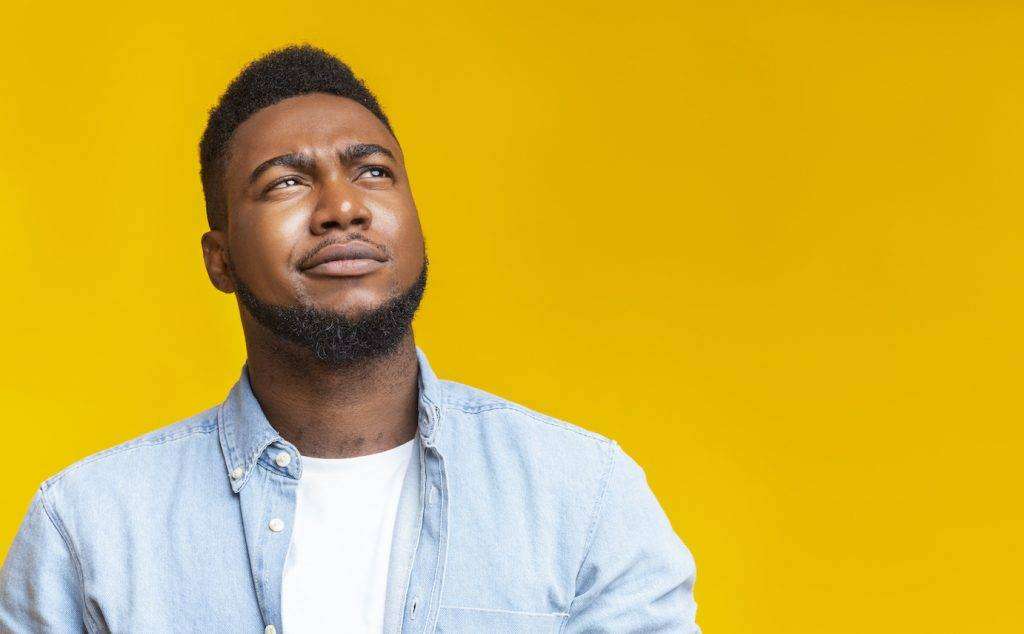 Portrait of suspicious black guy looking upwards at copy space on yellow background.